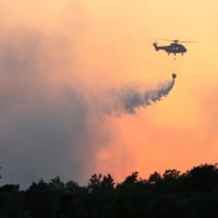 A helicopter silhouetted above a smokey forest against a burnt orange sky