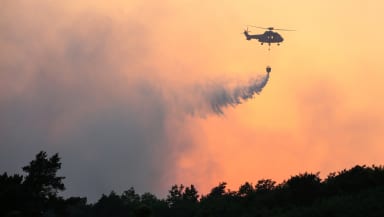 A helicopter silhouetted above a smokey forest against a burnt orange sky
