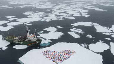 A Greenpeace ship alongside and ice floe that's covered in the flags of the world arranged in a heart shape.