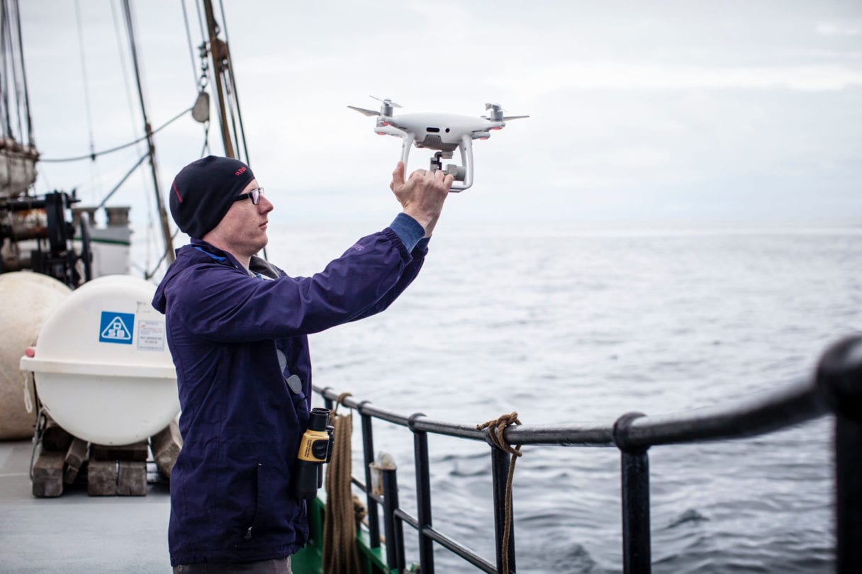 A man standing on the deck of a ship holds a drone above his head, preparing to launch it.