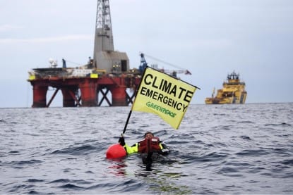 A swimmer floats in the sea, a short distance away from an oil rig. The flag they hold says "Climate emergency"