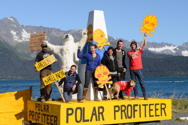 Campaigners stand on a platform with mountains in the background. The platform is emblazoned with 'Polar profiteer'. They hold signs saying "Shell no!" and shaped like a skull and cross in yellow and red.