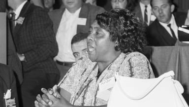 Fannie Lou Hammer is a Black woman. Her mouth is open mid-speech, as she sits and commands the attention of the people surrounding her.