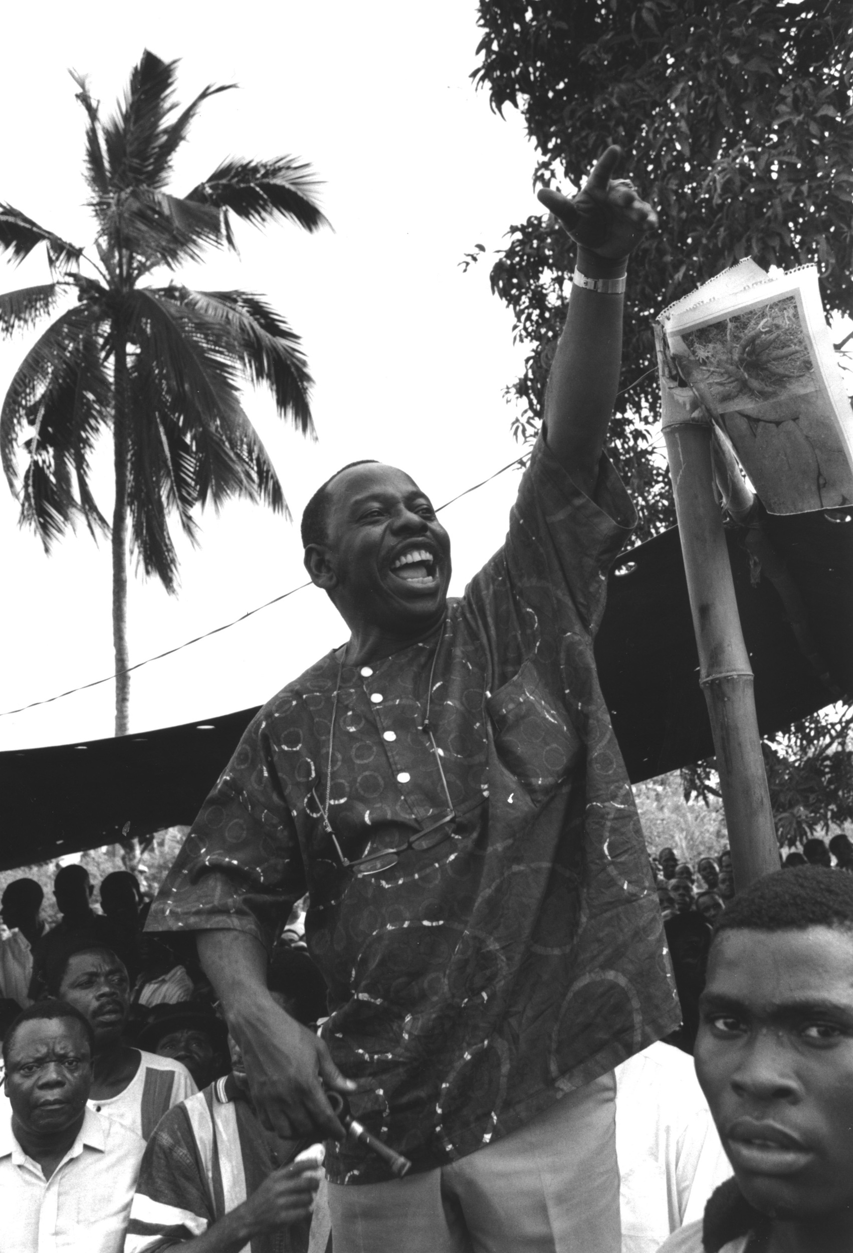 Monochrome photo of a Black man elevated slightly above a large crowd. He's jubilantly raising one hand in the air, with his index finger pointed aloft. 