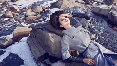 A model in a fashionably long grey coat lies on a rock in the middle of a polluted river
