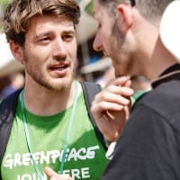 A fundraiser in a Greenpeace t-shirt talks to a member of the public in bright sunshine.