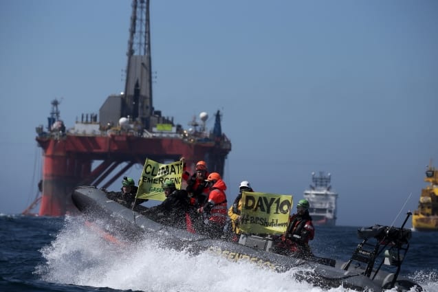A Greenpeace inflatable boat splashes over a wave. On board, activists hold up climate emergency banners. A giant oil platform can be seen in the background.