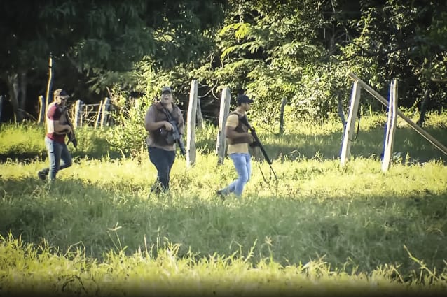 Three men armed with rifles walk through a field alongside a fence.
