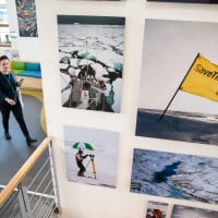 A staff member walks into the Greenpeace office. Colourful seating and large wall-mounted campaign photos can be seen in the background.