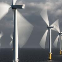 Offshore wind turbines lit by sun against a grey sky. Their blades are motion blurred.