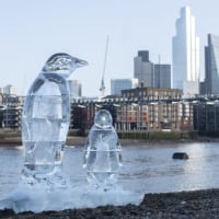Two ice scuplture penguins melt on the banks of the River Thames with the city and Millennium footbridge in the background