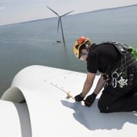 A worker in a hard hat and harness crouches on top of an offshore wind turbine. Other turbines are visible in the background.