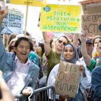 In bright sunshine, a smiling crowd of people hold up signs calling for a greener world. One protestor in the front row raises his fist in the air triumphantly.