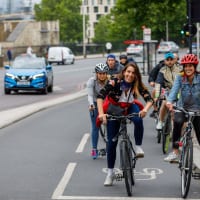 Two women on bikes smiling at the camera while waiting at a red light on a separated cycle lane. A queue of other riders is out of focus behind them.