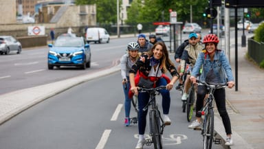 Two women on bikes smiling at the camera while waiting at a red light on a separated cycle lane. A queue of other riders is out of focus behind them.