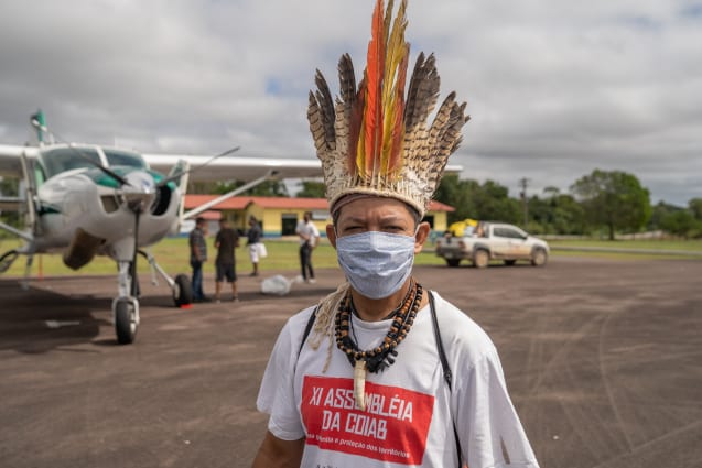 Person in a traditional Indigenous headdress and facemask stand in front of a light aeroplane on a runway