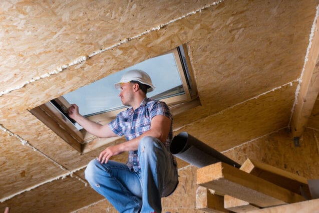 A worker in a hard hat adjusts a skylight window in a chipboard-lined loft space