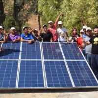 A group of trainees gathered around a small floor-mounted solar array. They're posing and smiling into the camera