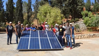 A group of people gathered around a small ground-mounted solar array, smiling at the camera.