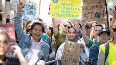 In bright sunshine, a smiling crowd of people hold up signs calling for a greener world. One protestor in the front row raises his fist in the air triumphantly.