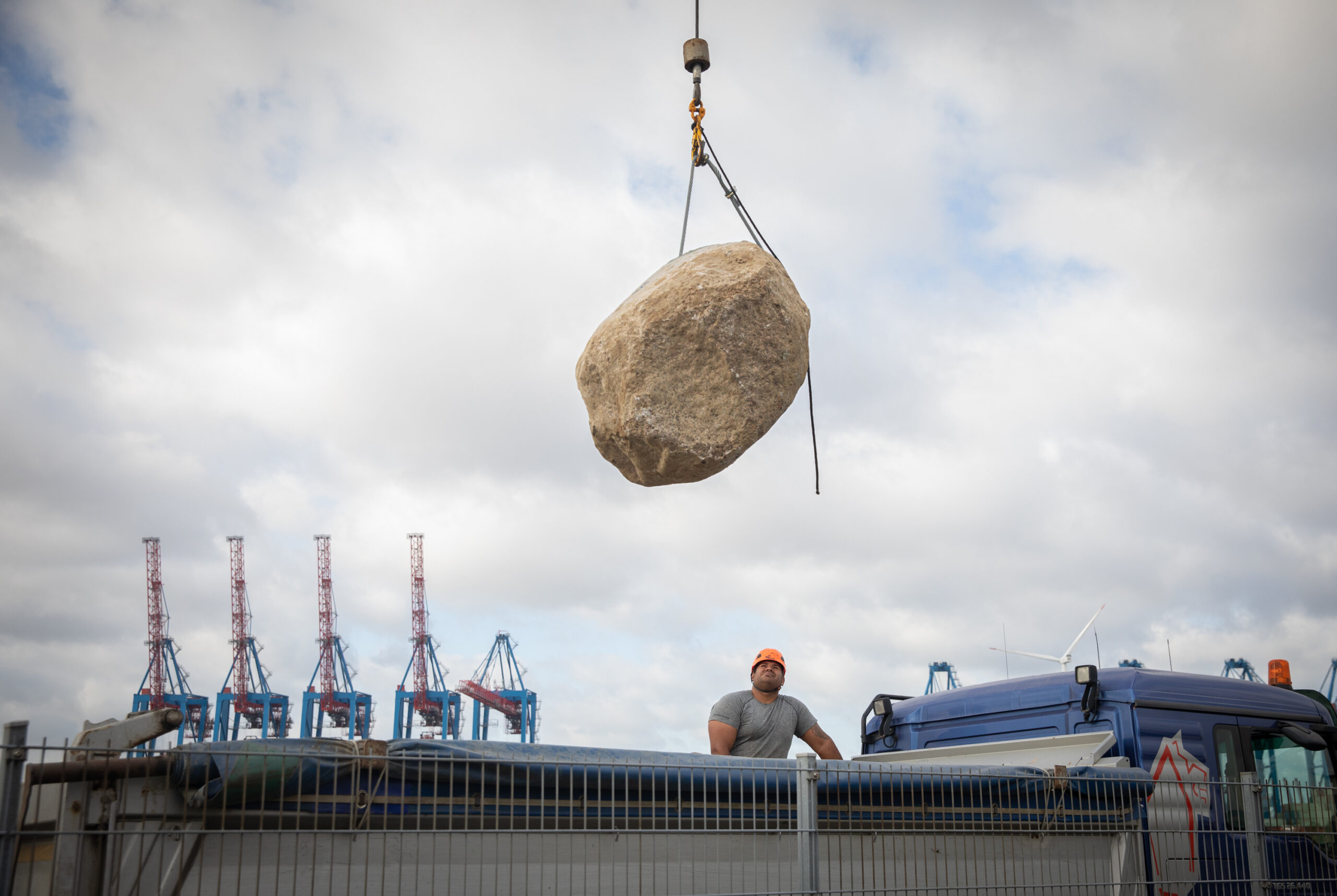 A Greenpeace crew member looks on as a boulder is lifted on board the ship. A row of giant port cranes are visible in the background