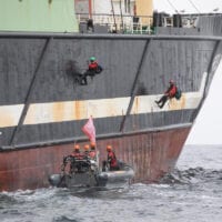 Two activists hang from the side of a large fishing ship. A small Greenpeace boat drives alongside.