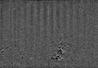Grey sonar of sea floor with visible comb-like lines. In the bottom centre-right a few impressions of the shipwreck are visible, with some debris scattered nearby