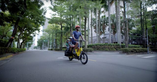 A parent carries two children on a cargo bike along an urban road