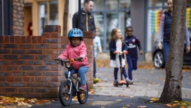 A young boy rides a bike along a residential street. A young girl on a scooter, a young boy running, and an adult walking are visible in the background.