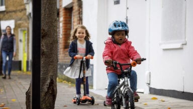 A young child on a bike rides towards the camera with a look of concentration on their face. Another child rides behind on a scooter.