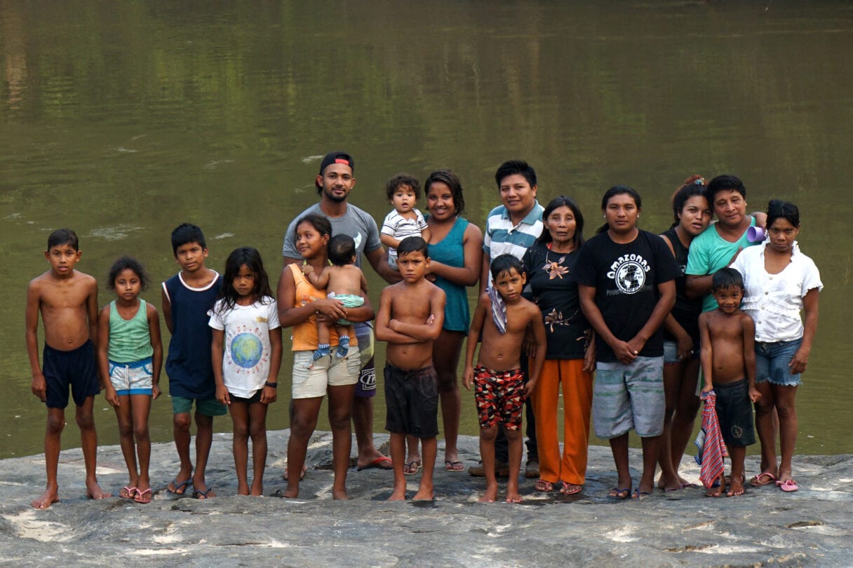 A group of around 20 adults and children stand by a river in a group, all looking into the camera. Some of the children are wet from swimming in the river.