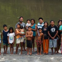 A group of around 20 adults and children stand by a river in a group, all looking into the camera. Some of the children are wet from swimming in the river.