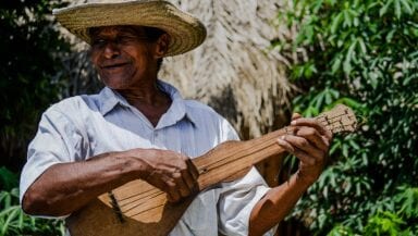 A man in a white shirt and straw hat smiles as he plays a traditional wooden string instrument