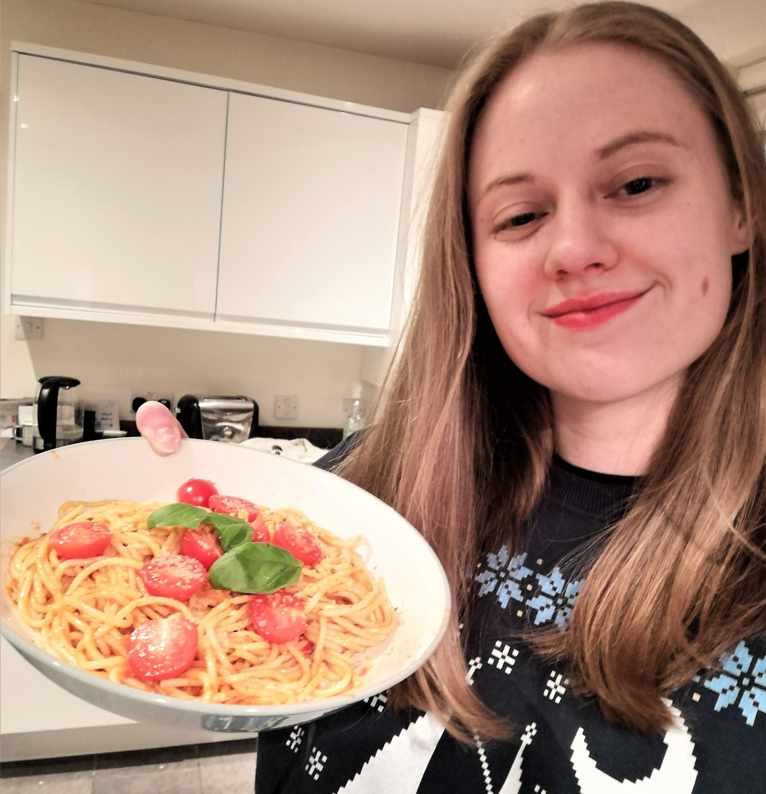 Kirsty smiles and holds up a plate of pasta decorated with cherry tomatoes and basil leaves. She's wearing a Greenpeace Christmas jumper
