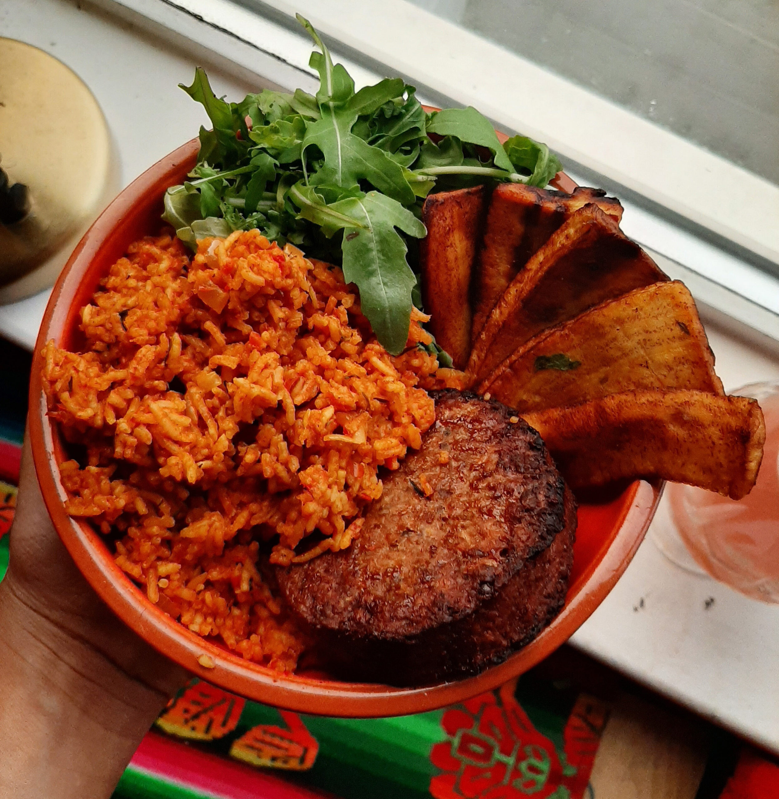 Overhead view of a bowl of jollof rice with a veggie burger, sliced plantain and rocket leaves arranged attractively