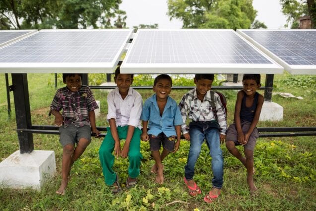 Children sit on the frame underneath a row of solar panels, smiling at the camera.