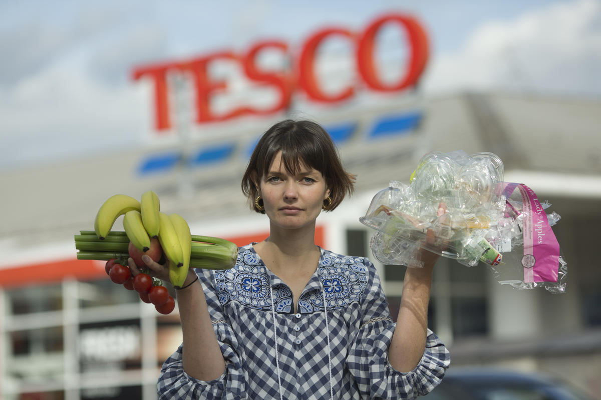 A person stands outside a Tesco superstore holding up fruit and vegetables in one hand and plastic packaging in the other.