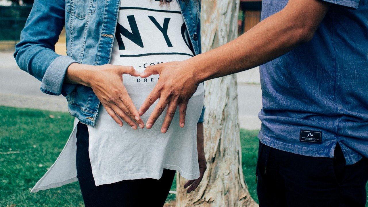 A couple's hands combine to make a heart shape over a woman's baby bump. She wears a denim jacket and an oversized white tshirt