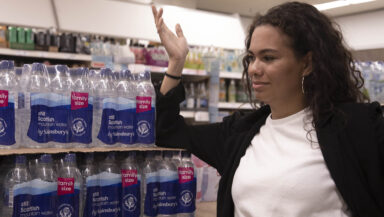 A shopper makes an exasperated gesture towards a head-high stack of plastic-wrapped bottled water multipacks