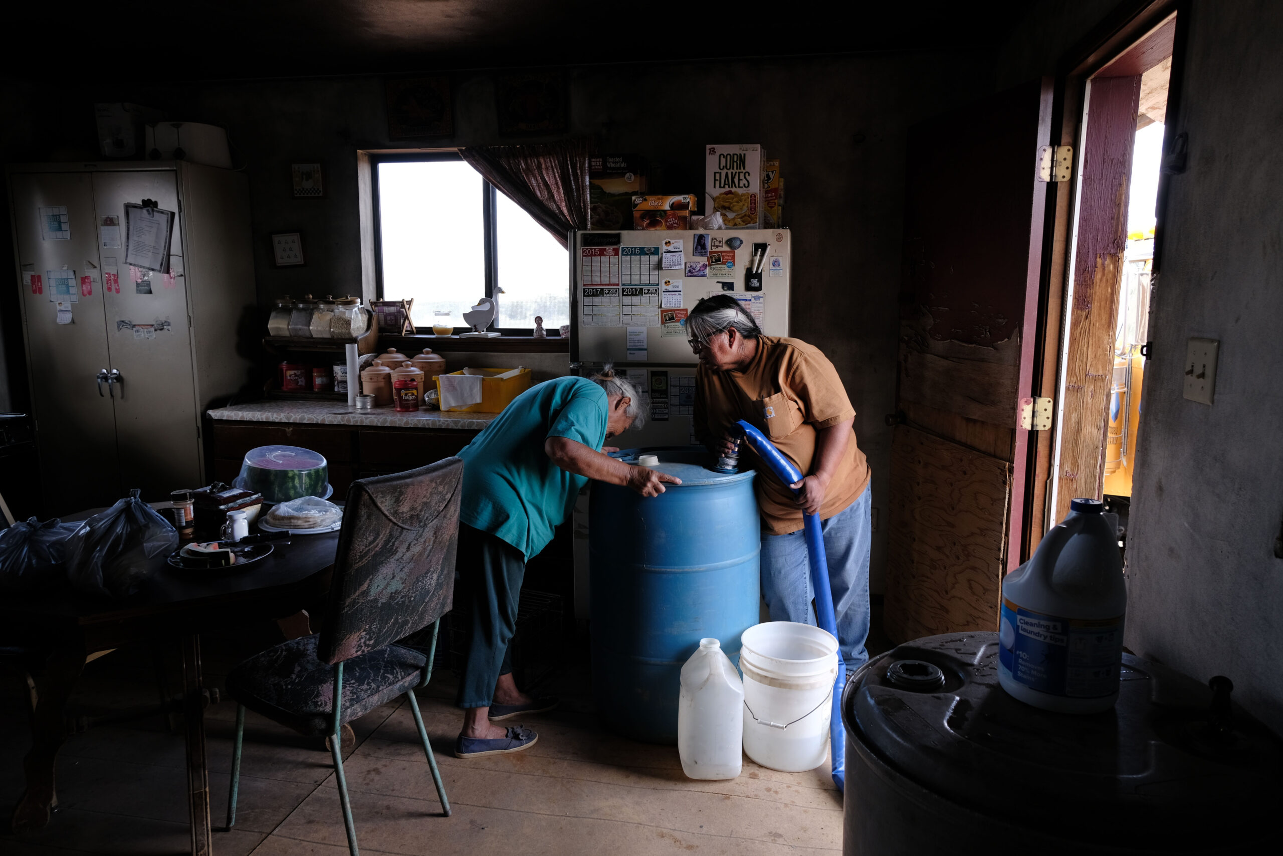 Indoor scene in a kitchen with a two older Navajo women and a large blue water tank with bottles and buckets beside it. The house is small and dark and cluttered.