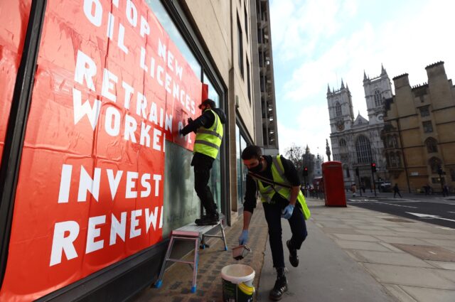 Two activists in hi-vis jackets paste a large red and white poster onto a window on a city street. A cathedral is visible in the background.