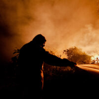 A firefighter sprays water on a blaze, silhouetted against a background of orange smoke