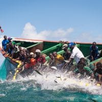 A dozen or so small-scale fishermen in a colourful wooden boat pull up a net from a turquoise ocean.