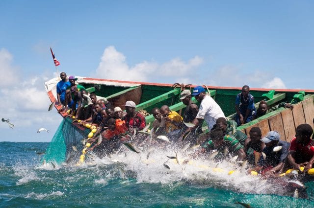 A dozen or so small-scale fishermen in a colourful wooden boat pull up a net from a turquoise ocean.