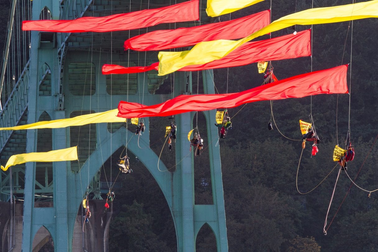 Activists in climbing gear hang from a giant river bridge. Huge red and yellow flags attached to their ropes fly proudly in the wind.