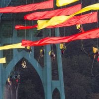 Activists in climbing gear hang from a giant river bridge. Huge red and yellow flags attached to their ropes fly proudly in the wind.
