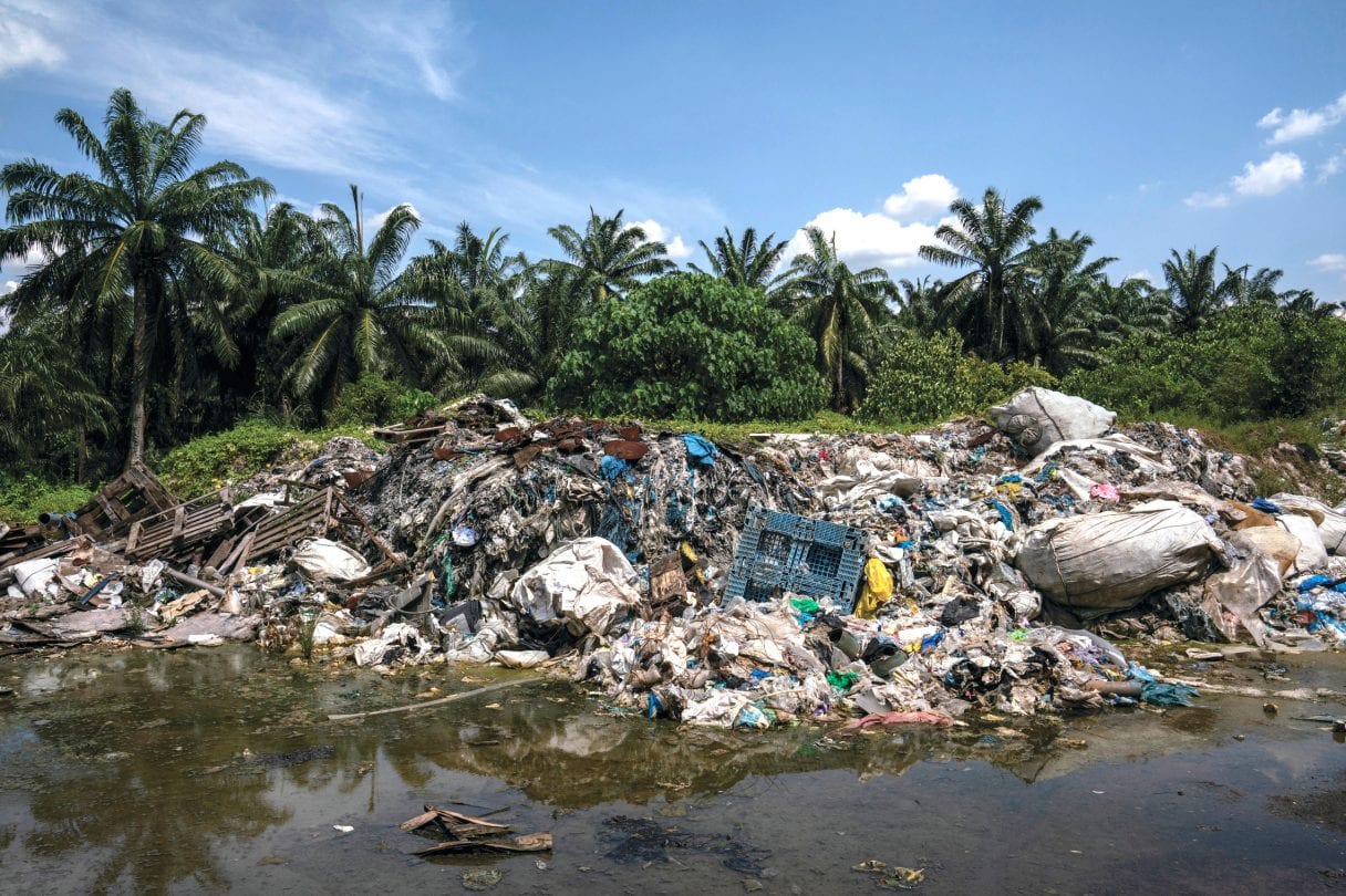 A pile of plastic waste sits next to a lake or river, with palm trees in the background.