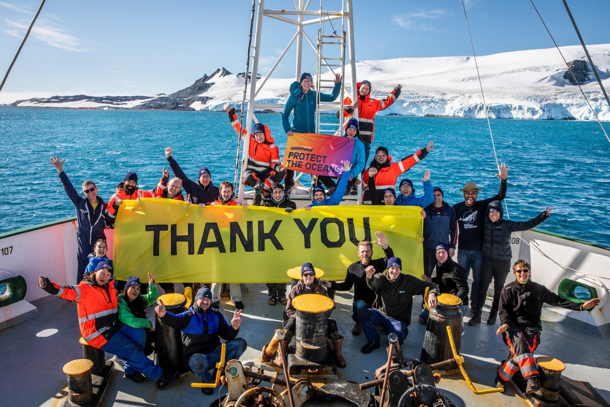 Large group on board the deck of a ship in a snowy environment wave to the camera and hold up a banner reading 'Thank you!'