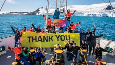 Large group on board the deck of a ship in a snowy environment wave to the camera and hold up a banner reading 'Thank you!'