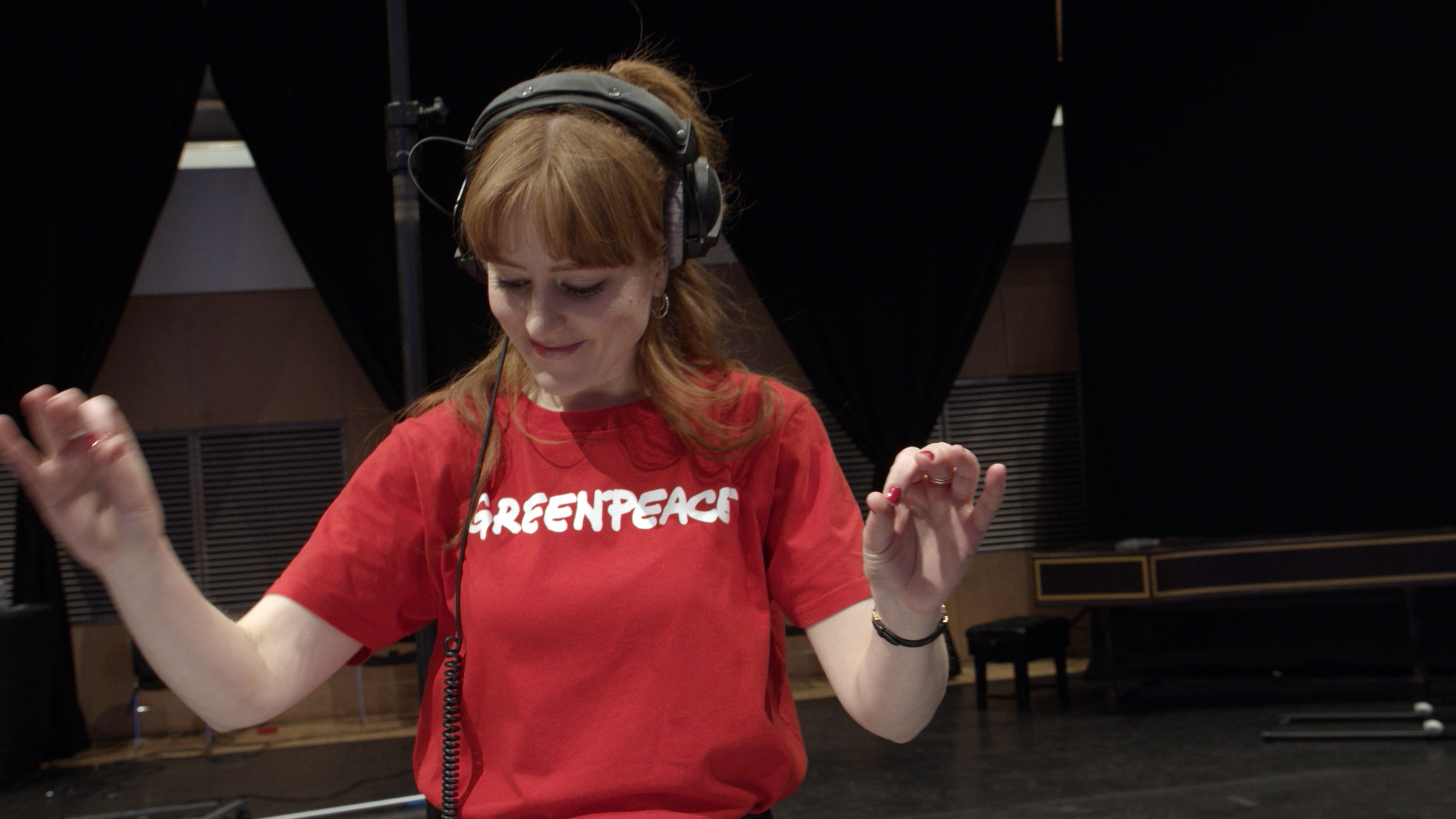 A conductor wearing headphones and a red Greenpeace tshirt looks down and smiles as she conducts an Orchestra